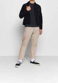 Svart mockajacka, grå tröja, beige byxor, vita strumpor och svarta sneakers. Jackan har två bröstfickor och framknäppning.