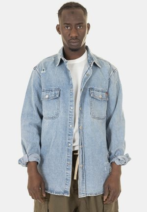 Camicia di denim azzurro chiaro con chiusura a bottoni, due tasche sul petto, dettagli strappati e maniche arrotolate, indossata sopra una t-shirt bianca.
