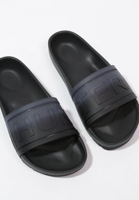 Une paire de sandales noires à enfiler avec des semelles texturées et de larges sangles embossées du mot "HUNTER" sur fond blanc.