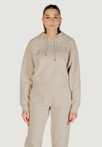 Felpa beige con cappuccio, caratterizzata da un logo "GUESS" scintillante, realizzata in morbido tessuto con vestibilità comoda, cordini regolabili e polsini a costine.