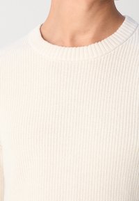 Cremefarbener, gerippter Pullover mit rundem Halsausschnitt und strukturiertem Strickmuster. Der Stoff wirkt weich und leicht elastisch.