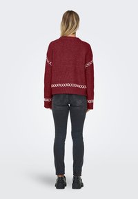Blond kvinne står med ryggen til iført en vinrød genser med hvite korsstingdetaljer, mørke tettsittende jeans og svarte platåsko.