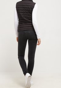 Svart pufferväst med horisontellt quiltat mönster, bärs över en vit långärmad skjorta, ihop med åtsittande svarta jeans och vita sneakers.
