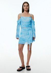 Himmelblaues, transparentes Kleid mit quadratischem Ausschnitt, schulterfreien langen Ärmeln und Raffung. Mit einem Knoten-Detail an der Taille, kombiniert mit Sandalen.