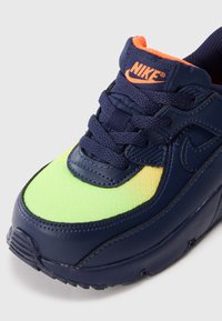 Nikesneaky s modrými šnúrkami, podrážkou a prekrytiami, neonovo zeleno-žltým špičkovým boxom a oranžovým logom na jazyku na bielom pozadí.