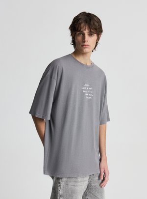 Młody mężczyzna o brązowych włosach, ubrany w luźny szary T-shirt z białym nadrukiem oraz jasnoszare, wyprane jeansy, stoi na tle gładkiego, jasnego tła.