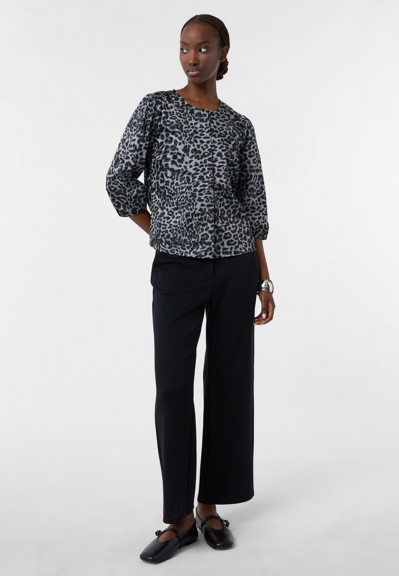 Schwarze und graue Leopardenmuster-Bluse mit puffärmel, kombiniert mit hochgeschnittenen schwarzen Hosen und flachen schwarzen Schuhen. Glatte Textur.