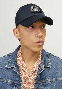 Gorra de béisbol azul marino con un logotipo en parche, combinada con una camisa estampada debajo de una chaqueta de mezclilla. Presenta un ala curvada y parte trasera de malla.