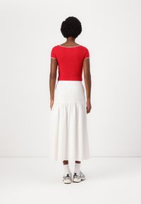 Top corto rojo de manga corta con un cuello redondeado, combinado con una falda larga blanca con volantes. La modelo lleva zapatillas plateadas y calcetines blancos.