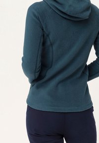 Teal fleece hoodie with a hood, smooth texture, side seams, and a rounded hem. Paired with dark pants, viewed from the back.