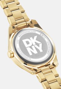 Orologio in acciaio inox dorato con quadrante grigio. Presenta un grande logo "DKNY", resistenza all'acqua fino a 5 ATM e un design del bracciale texturizzato.