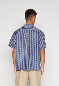 Košile s krátkými rukávy s modro-bílým geometrickým vzorem, vyrobená z lehkého materiálu s špičatým límečkem a detaily na zadním díle.