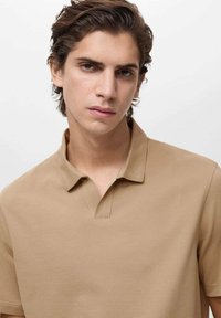 Beżowa koszulka polo z kołnierzykiem, teksturowany materiał i czysty design. Nosi ją model na neutralnym tle w naturalnym oświetleniu.