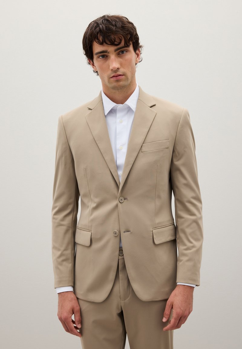 Giacca da suit beige in tessuto liscio con una tasca sul petto, due tasche frontali e chiusura a due bottoni; abbinata a una camicia bianca.
