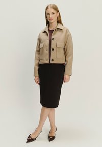 Giacca cropped in lana beige con due tasche anteriori e bottoni neri, indossata sopra un top bordeaux, abbinata a una gonna a tubino nera aderente e tacchi scuri.