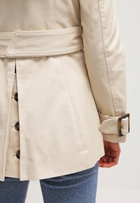 Trench-coat beige avec une taille cintrée et une ceinture. Il présente de gros boutons marron et un tissu lisse et texturé.