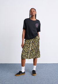 Svart oversized t-skjorte med grafisk trykk, kombinert med en leopardmønstret skjørt i grønn og brun som går til kneet og har glidelås foran, supplert med svarte sneakers.
