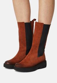 Brązowe zamszowe botki za kostkę z czarnymi elastycznymi panelami bocznymi i zaokrąglonym noskiem, wyposażone w grubą czarną podeszwę dla dodatkowego komfortu.