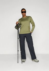 Oliiviroheline pika varrukaga särk taskuga, kantud tumedanavüürist pükstega. Modell hoiab matkakeppe ja kannab halli matkajalatsit.