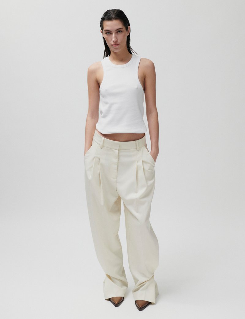 Femme aux cheveux mouillés portant un haut blanc ajusté sans manches, un pantalon large crème taille haute et des chaussures marron pointues, debout devant un fond uni.