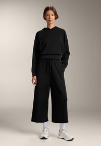 Zwarte oversized hoodie met een voorzak en een trekkoord. Gecombineerd met wijde zwarte sweatpants. Kenmerkt witte sportschoenen en geribbelde sokken.