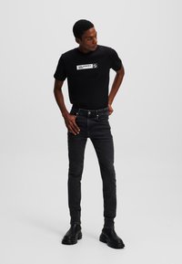 Camiseta negra con logo blanco, combinada con jeans negros ajustados. Modelo con botas negras gruesas, de pie en un fondo neutro.