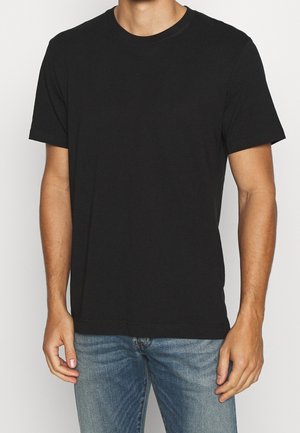 Homme portant un t-shirt noir uni à manches courtes et col rond, et un jean bleu, debout devant un fond clair uni.