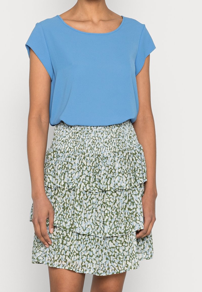 Femme portant un haut bleu à manches courtes rentré dans une mini-jupe à volants verte et bleu clair à motifs, avec une ceinture froncée.