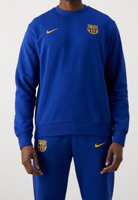 Conjunto de sudadera y pantalones azules, confeccionado con tela suave. Presenta el logo de Nike y el emblema del FC Barcelona en amarillo, con un diseño de cuello redondo.