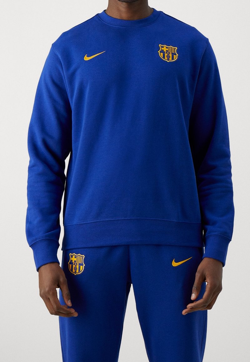 Conjunto de sudadera y pantalones azules, confeccionado con tela suave. Presenta el logo de Nike y el emblema del FC Barcelona en amarillo, con un diseño de cuello redondo.