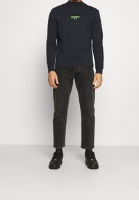 Man som bär en svart höghalsad långärmad tröja med en liten grön och vit logotyp, svarta jeans, svarta strumpor och svarta sneakers, stående upprätt.