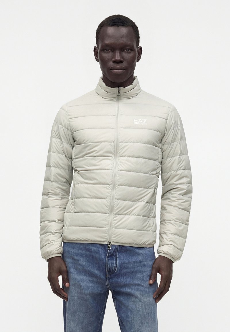 Homme portant une veste matelassée gris clair avec logo EA7 sur la poitrine et un jean bleu, debout devant un fond blanc uni.