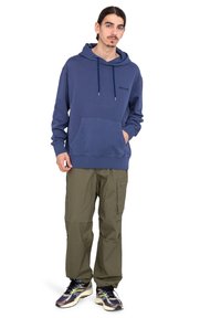 Sudadera azul marino con bolsillo frontal y capucha ajustable, combinada con pantalones cargo en color verde oliva y zapatillas multicolores.