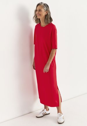 Smilende kvinde med gråt hår iført en rød midi-kjole med sidespalte og hvide sneakers, der står mod en hvid mur.