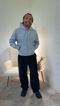 Νέος άνδρας που χαμογελά με τα χέρια στις τσέπες, φοράει γαλάζιο φούτερ με κουκούλα, μαύρο παντελόνι και μαύρα παπούτσια, στέκεται σε ένα δωμάτιο με καρέκλα και μικρό τραπέζι.