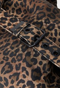 Tecido com padrão de leopardo em tons de castanho e preto, apresentando uma fivela preta robusta e rebites. Superfície texturizada e detalhes de costura visíveis.