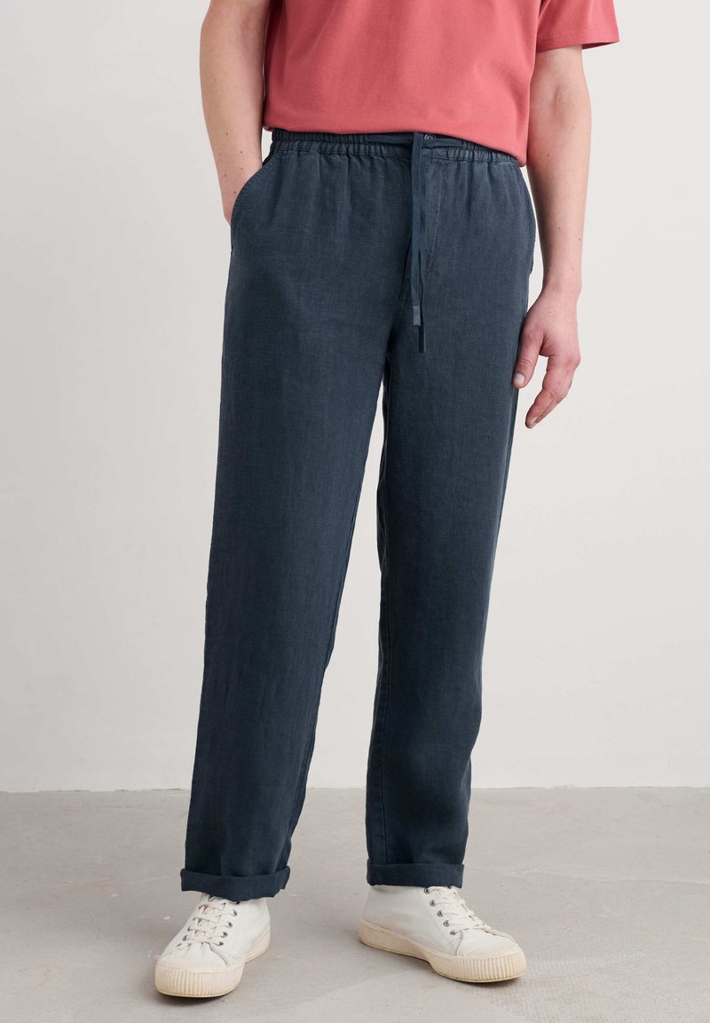 Navy blue linen pants with an elastic waistband and drawstring, featuring side pockets and rolled cuffs, paired with white sneakers.