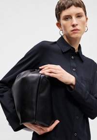 Črna usnjena torba pravokotne oblike z gladko teksturo. Model nosi temno srajco z gumbi in dolgimi rokavi, drži torbo.