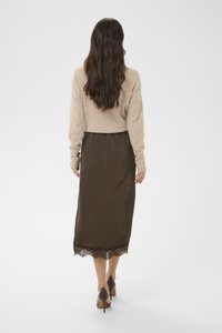 Femme aux longs cheveux bruns ondulés portant un pull beige, une jupe midi marron à bord en dentelle et des chaussures à talons marron, debout de dos.