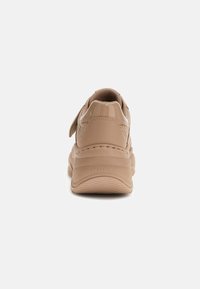 Beżowe, masywne sneakersy z grubą podeszwą, z rzepem i wytłoczonym logiem "GUESS" na pięcie, pokazane z tyłu na białym tle.