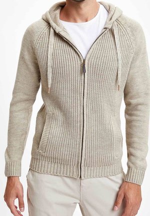 Mand iført en beige strikket lynlåshættejakke med forlommer og en hvid t-shirt under.