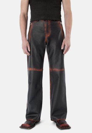 Personne portant un pantalon en cuir noir avec des accents orange usés, des fermetures éclair près des hanches, associé à des chaussures à bout carré et un haut noir sans manches.