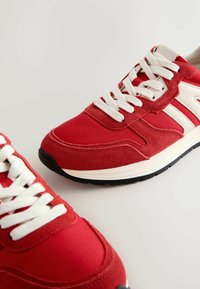 Adidași roșii realizati dintr-o combinație de pânză și piele întoarsă, cu șireti albi, guler căptușit și talpă din cauciuc neagră.