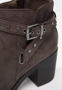 Gros plan sur une botte cheville en suède gris foncé avec boucle argentée, détails de sangle et talon empilé noir épais.