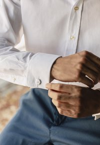 Camicia bianca con gemello esagonale. Il tessuto appare liscio con una leggera lucentezza. I polsini sono abbottonati, con dettagli di cucitura sottili.