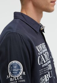 Navyfarbendes Langarmhemd mit Knopfkragen, ausgestattet mit weißem Drucktext und einem runden blauen Patch am linken Ärmel. Strukturiertem Stoff.