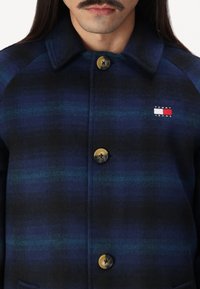 Jakna od vune s kariranim uzorkom u tamnoplavoj boji, s zelenim prugama, oštrim kragnom i zlatnim dugmetima. Mali Tommy Jeans logo na grudima.