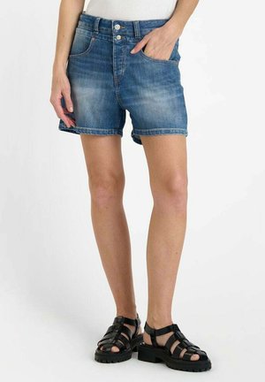 Shorts en denim bleu clair avec une finition légèrement délavée, ourlet roulé, cinq poches et une fermeture éclair, associés à des sandales noires à lanières.