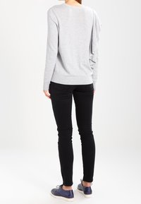 Grauer Langarmpullover mit Rüschendetail hinten, kombiniert mit schwarzen enganliegenden Hosen und blauen Schuhen. Schlichtes Design, weiche Textur.