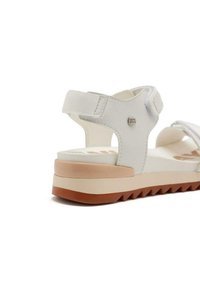 Sandalia blanca con tira ajustable, material texturizado, tacón curvado y una suela bicolor que presenta una base de goma marrón y una entresuela blanca.